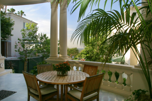 A serene outdoor patio with a wooden table, chairs, and lush greenery, framed by elegant columns and a clear sky.