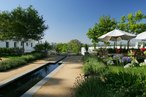 A serene garden with a water feature, lush greenery, and shaded seating under umbrellas against a clear blue sky.