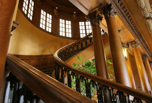 A grand wooden staircase spirals upward, flanked by ornate columns and large windows, creating an elegant interior.