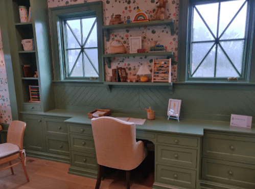 A cozy green desk area with shelves, a chair, and decorative items, illuminated by natural light from two windows.