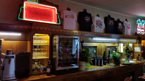A cozy restaurant bar with a neon sign, shirts on display, and shelves of drinks and plants.