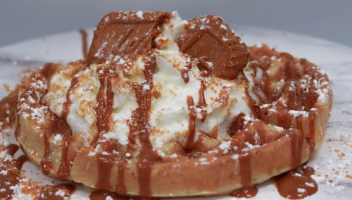 A delicious waffle topped with whipped cream, caramel drizzle, and a cookie, sprinkled with crumbs.