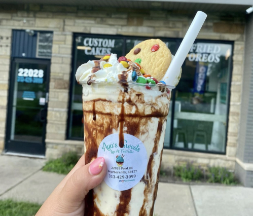 A hand holds a large milkshake topped with whipped cream, chocolate drizzle, sprinkles, and a cookie, with a shop in the background.