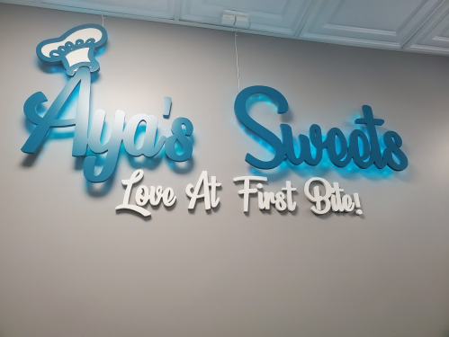 Sign for "Aya's Sweets" with the tagline "Love At First Bite!" in blue and white lettering on a light wall.