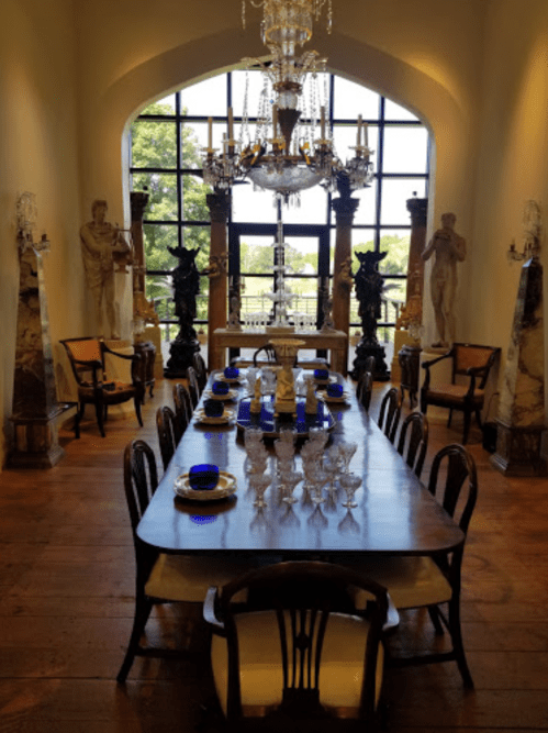 Elegant dining room featuring a long wooden table, ornate chandelier, and classical sculptures in a bright, spacious setting.