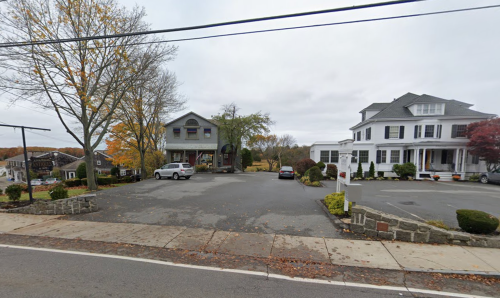 A quiet street scene featuring two large houses, a gravel parking area, and autumn trees under a cloudy sky.