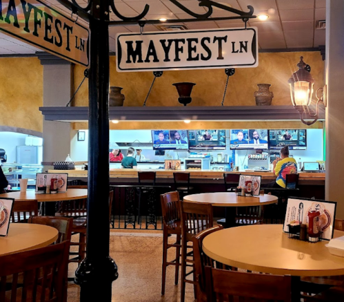 Interior of a restaurant with tables, a bar area, and signs for "Mayfest Ln," featuring a warm, inviting atmosphere.