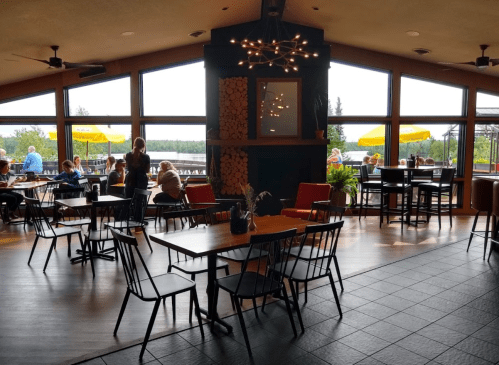 Interior of a restaurant with large windows, tables, and chairs, overlooking a scenic view with patrons dining.