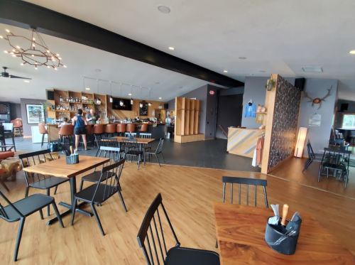 A modern restaurant interior with wooden tables, black chairs, a bar area, and a warm, inviting atmosphere.