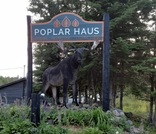 Sign for Poplar Haus featuring a moose statue, surrounded by trees and greenery, indicating lodging, restaurant, and liquor store.
