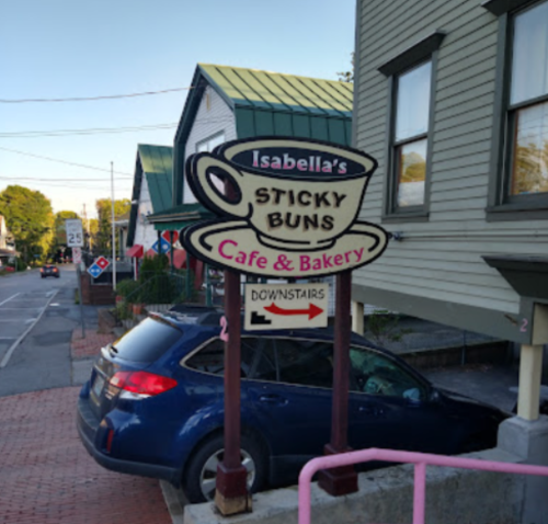 Sign for "Isabella's Sticky Buns Cafe & Bakery" with a coffee cup graphic, located on a street with houses nearby.