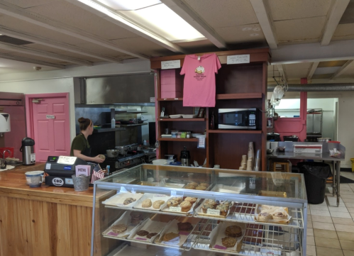 A cozy kitchen with pink accents, featuring a worker at the stove and a display case of baked goods.