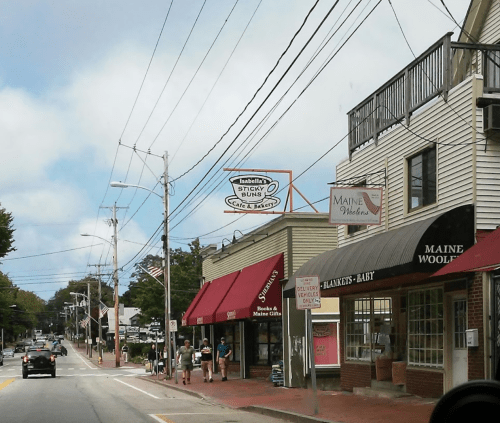 A street view of a small town with shops, signs, and people walking along the sidewalk under a cloudy sky.