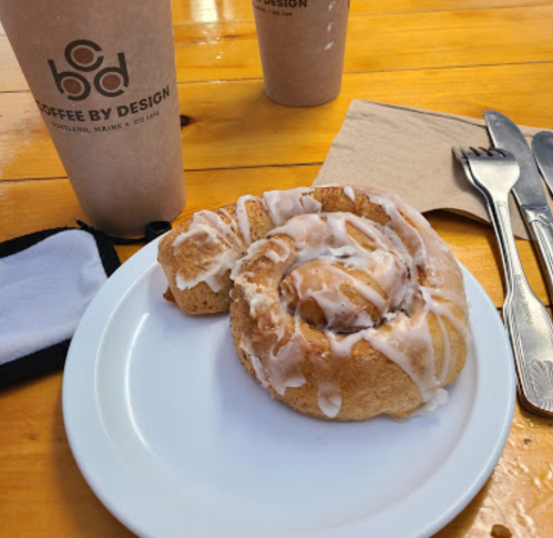 A cinnamon roll with icing on a white plate, accompanied by coffee cups and utensils on a wooden table.