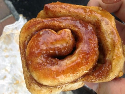 A close-up of a cinnamon roll with a glossy glaze, held in a hand, showcasing its swirled layers.