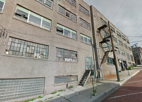 A gray industrial building with large windows and a metal fire escape, viewed from the street.