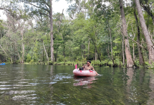A person relaxes on a pink inner tube in a serene, green river surrounded by trees.