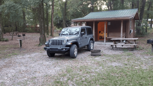 A gray Jeep parked near a cabin in a wooded campsite with picnic tables and a fire pit.
