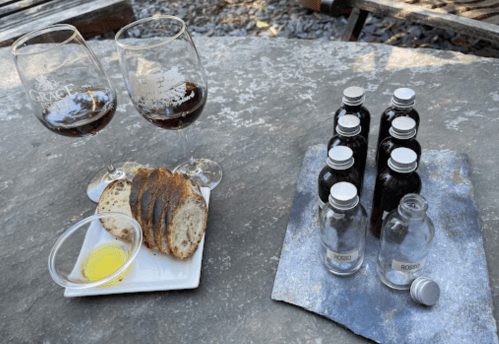 Two glasses of wine beside a plate of bread with olive oil, and a tray of small bottles labeled "Rosso."