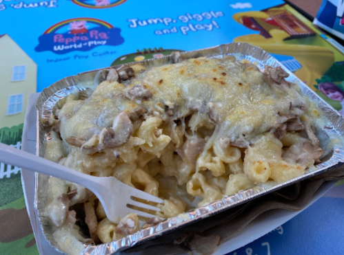 A cheesy pasta dish with chicken, served in a foil container, on a colorful Peppa Pig-themed table.