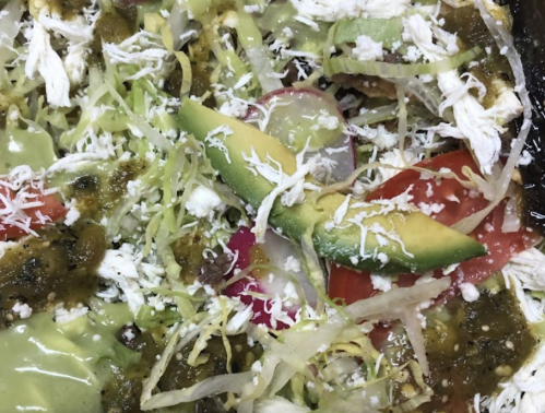 A close-up of a colorful dish featuring shredded chicken, lettuce, tomatoes, avocado, and cheese, drizzled with green sauce.