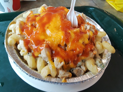 A foil bowl filled with macaroni, chicken, and melted cheese, topped with a drizzle of sauce and a plastic fork.