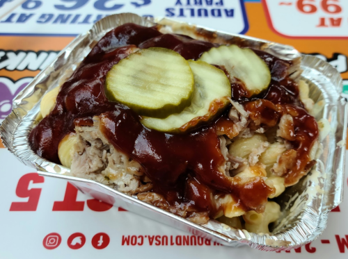 A foil container filled with macaroni, pulled pork, barbecue sauce, and pickles on top.