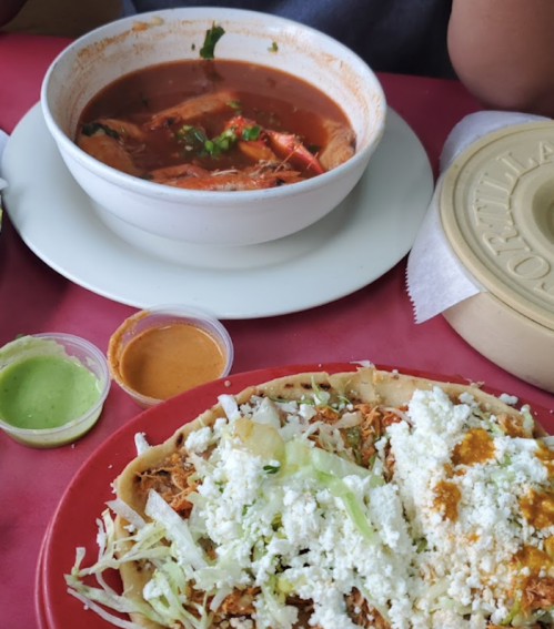 A bowl of soup with herbs and vegetables next to a plate of tacos topped with lettuce and cheese, with sauces on the side.