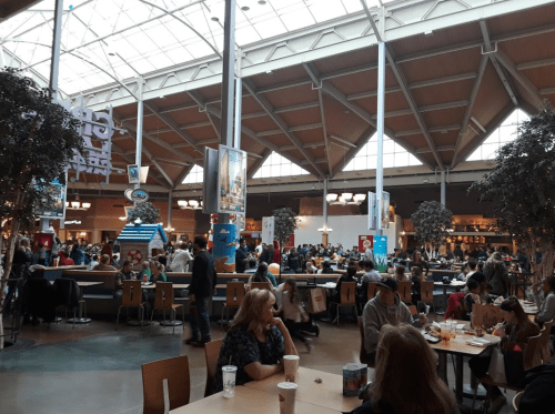 A busy food court filled with people dining at tables under a large, bright skylight.
