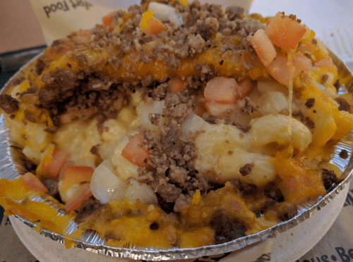 A close-up of a cheesy, meaty dish topped with diced tomatoes and crumbled meat in a foil container.