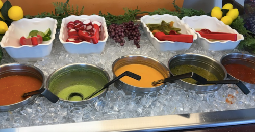 A colorful display of sauces and toppings in bowls on ice, surrounded by fresh fruits and vegetables.