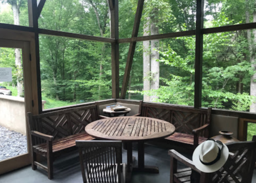 A cozy screened porch with wooden benches and a round table, surrounded by lush green trees.