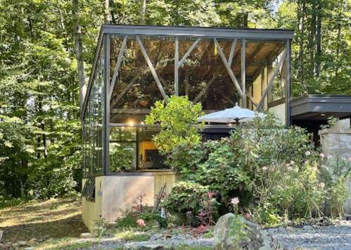 A modern glass-walled house surrounded by greenery and flowers, featuring a patio umbrella and natural light.