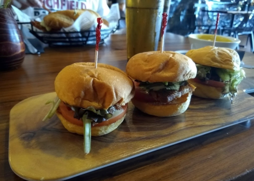Three mini burgers on a wooden platter, topped with lettuce, tomato, and skewered with red picks.