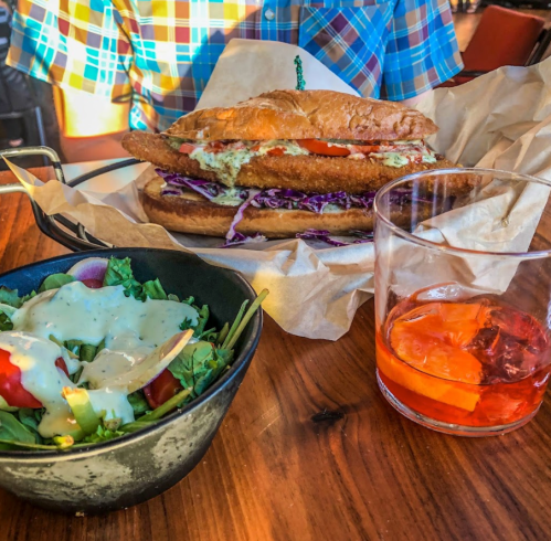 A large sandwich with fried filling, slaw, and tomato, served with a side salad and a drink on a wooden table.