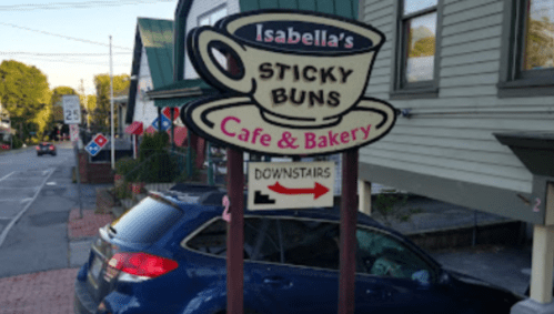 Sign for Isabella's Sticky Buns Cafe & Bakery, featuring a coffee cup design, with a blue car parked nearby.