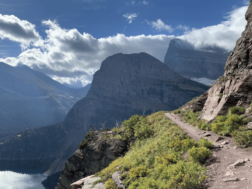 A scenic mountain trail with rocky cliffs, lush greenery, and a lake under a partly cloudy sky.