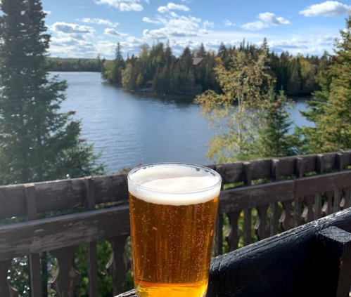 A glass of beer sits on a railing, overlooking a serene lake surrounded by trees and a clear blue sky.