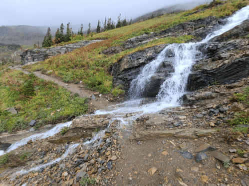 A rocky hillside with a small waterfall cascading down, surrounded by green grass and trees under a cloudy sky.