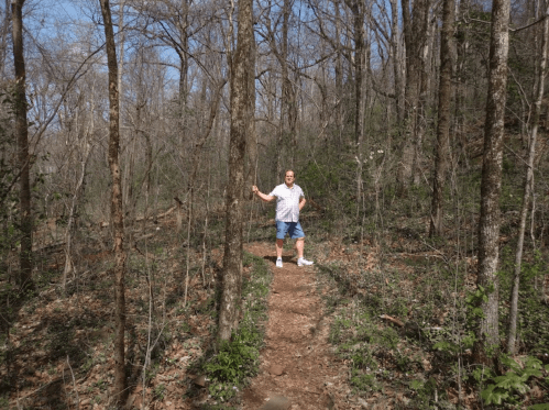 A person stands on a dirt path in a wooded area, surrounded by trees and greenery on a sunny day.