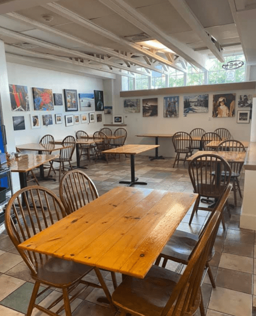 A cozy café interior with wooden tables and chairs, featuring art on the walls and large windows for natural light.