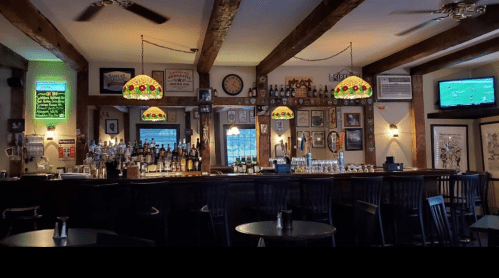 A cozy bar interior with wooden beams, stained glass lamps, and a well-stocked bar, featuring tables and wall decor.