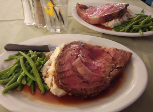 A plate of prime rib with mashed potatoes and green beans, served with a glass of water in the background.