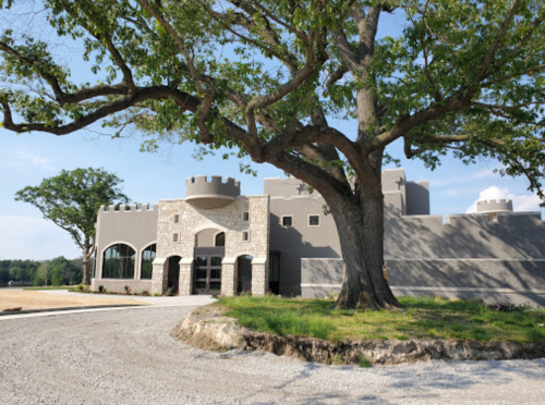 A large stone building resembling a castle, set beside a sprawling tree and gravel driveway under a clear blue sky.