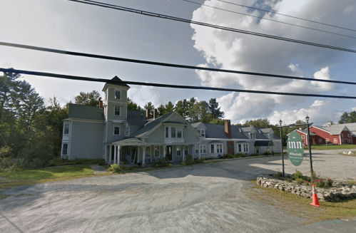 A quaint inn with a tower, surrounded by trees and a gravel parking area, under a cloudy sky.