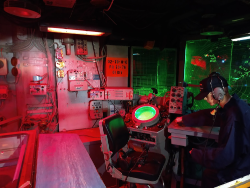 A dimly lit control room with red and green lights, featuring various equipment and a person seated at a desk.