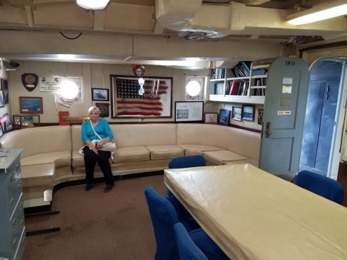 A woman sits on a beige couch in a room with nautical decor, including an American flag and various framed pictures.