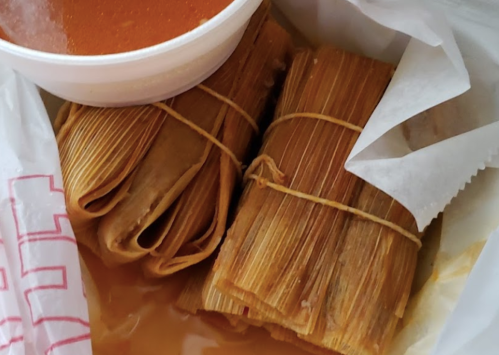 Two bundles of tamales tied with string, accompanied by a small bowl of red sauce, in a takeout container.