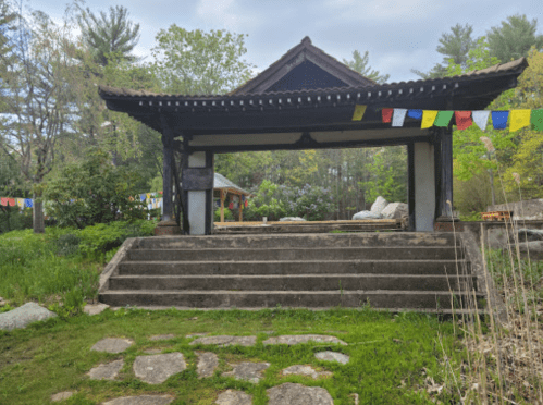 A serene entrance with stone steps leading to a traditional structure, surrounded by greenery and colorful prayer flags.