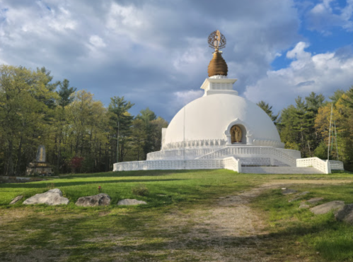 A white stupa surrounded by trees under a cloudy sky, with a statue visible nearby on a grassy path.